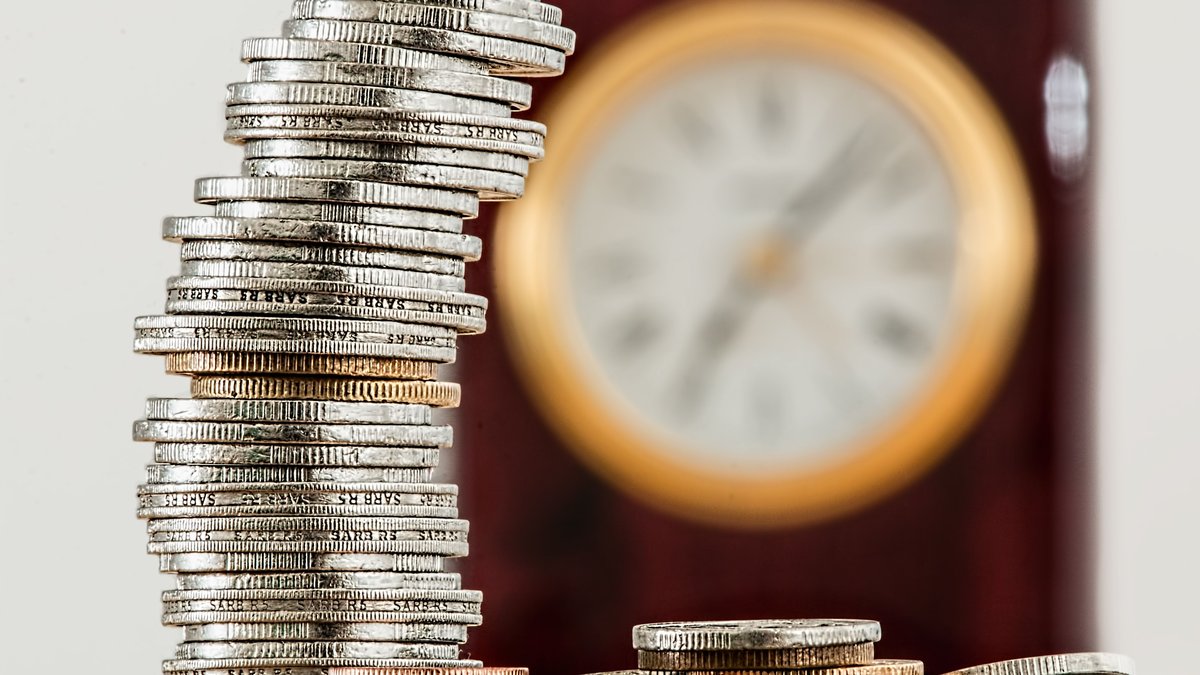 Pilha de moedas em foco com relogio ao fundo, simbolizando o tempo no juros compostos